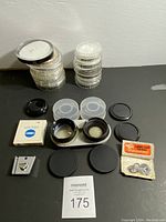 Full lot overview showing stacked circular filter cases, black lens caps, Minolta filter box, two lens converters in holder, metal clip accessory, and lens cap leash