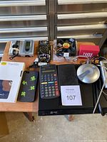 Photo showing various electronic items arranged on a table: calculator, thermometer parts, speaker box, computer fan, MP3 player, external device, phone case, and more.
