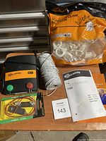 Gallagher electric fence energizer with terminals, spool of white wire, package of white plastic mounting brackets, electrical connectors and user manuals on wooden surface.