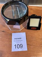 Photo of black lighted lupe and black KIMAC film viewer side by side on wooden surface with MaxSold auction card 109