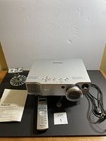 Panasonic projector PT-AE900U with power cord, mounting accessory, instruction manual, and remote control on black surface.