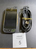 Front view of the SONIM XP-7700 phone alongside a charging cable, showing the device screen, buttons, and antenna.