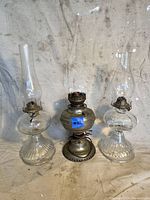 Full view of three antique oil lamps showing two ribbed glass bases with tall chimneys and one round glass font lamp with metal burner.