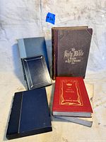 Six antique books arranged on a surface, showing a large Holy Bible book, Ideal Mother, and several other hardcovers with worn edges and slightly faded covers.