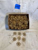 Top-down view of a box filled with approximately 166 brass flower-shaped jewelry parts, with seven sample pieces displayed in front of the box showing the intricate loop design.