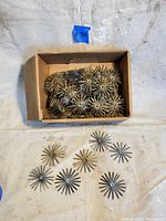 Top-down view of a cardboard box holding approximately 75 vintage brass wire jewelry findings with starburst design, some spread out outside the box for detail.