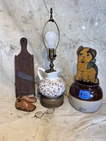 Full view of the lot including pottery jar, wooden dog figurine, ceramic pitcher lamp, wooden ribbed flatware holder, carved wooden duck, and eyeglasses.