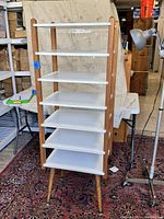 Front view of the shelving unit showing six white shelves, wooden vertical supports, and tapered wood legs with metal caps.