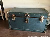 Photo of one metal steamer trunk closed on tabletop showing brass trim and lock latches, green exterior.