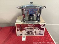 Stainless steel chafing dish on top of its original box, showing the front view with fuel burner visible inside the stand, and lid on the side.