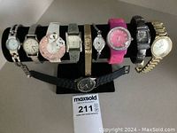 Front view of nine ladies fashion watches on display stand with MaxSold lot tag; showing various colors, designs, and band types including metal, silicone, and textured materials.