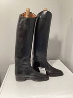 Front and side view of the pair of tall black leather riding boots standing upright on a white surface to show shape, height and condition.