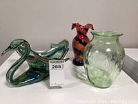 Front view of three pieces: green and blue swirled glass swan, green embossed animal motif vase, rust and blue swirl ruffled top vase.