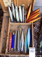 Top view of all 17 vintage heavy metal fishing lures arranged vertically inside a vintage wooden box. Different colors including metallic silver, orange with black spots, and blue with black spots are visible. Several lures have a scale pattern texture.