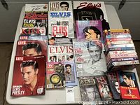 Flat lay of Elvis Presley magazines, books, VHS tapes, DVDs, and collectible records arranged on a table.