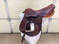 Side view showing full saddle with attached stirrup leathers and metal stirrups, saddle is dark brown leather with smooth finish.