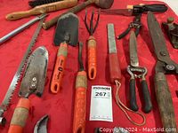 Photo of garden hand tools laid out on red cloth, including trowel, cultivator, hoes, pruning shears, saw, and other vintage hand tools.