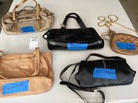 Top view of 5 handbags arranged on floor, showing various styles, colors, and brand tags labeled on blue tape.