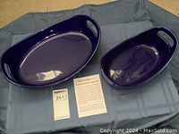 Top view of two oval cobalt blue stoneware baking dishes with handles placed on a blue cloth with MaxSold tags and Rachael Ray information card