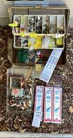 Contents of vintage fishing tackle box open to reveal multiple compartments with assorted small fishing lures with feathers, metallic finishes and bright colors; loose lures and packaged lures below the box