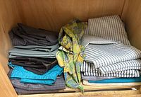 Photo of various barber capes folded and stacked on a wooden shelf, showing multiple colors and patterns.
