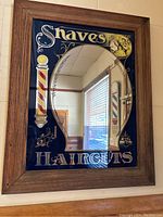 Front view of a vintage barber mirror with oval mirror center and painted dark glass border featuring barber poles and tools imagery with text 'Shaves and Haircuts'. Wood frame surrounding mirror.