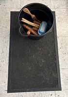 Photo of a large black rubber mat with a black plastic bucket containing multiple wooden handled brushes and a scrapper tool placed on top.