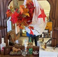Photo showing various collectible items on wooden surface in front of a mirror, including bottles, glassware, small train figurine, brass lamps, and decorative tins, with artificial autumn leaves hanging above.