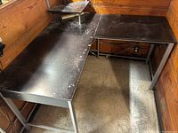 Photo showing top view of black L-shaped corner desk with worn surface, scratches and dust, set against wood paneled walls and linoleum floor, metal legs visible. Shows overall shape and condition.