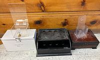 Three boxes side by side on floor against wood wall, showing white donation box with acrylic lock top, black metal box with holes, and wooden box with acrylic holder.