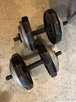 Two adjustable dumbbells on a floor, showing black and silver iron weight plates on threaded handles.