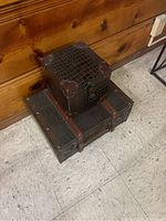 Two stacked vintage-style luggage boxes, one smaller with a crocodile texture and leather straps on top of a larger one with similar design, placed on a tiled floor against a wooden wall.