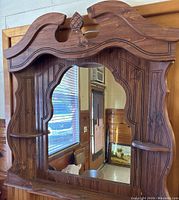 Front view of large wooden framed mirror with carved top and two small shelves.