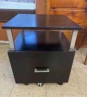 Front and side views of black printer stand file cabinet showing top surface, open shelf, bottom drawer with silver handle and caster wheels.