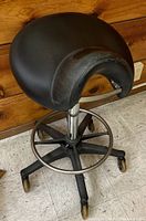 Photo of black rolling saddle stool showing padded seat, half backrest, adjustable metal base with footrest ring and caster wheels.