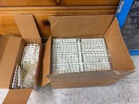 Two cardboard boxes containing multiple trays of white plastic tab size letters and numbers, placed on floor.