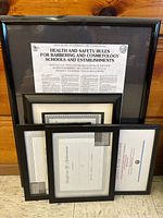 Five various sized black picture frames leaning against a wooden wall, each containing certificates or documents, in used condition and needing cleaning.