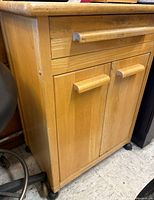Close-up showing one drawer above two cabinet doors with wooden handles, light wood finish, on wheels, scratches visible.