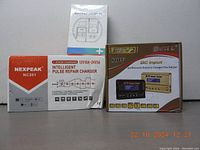 Photo showing the Nexpeak NC201 charger box and a small manual, alongside the HTRC B6V2 charger box in brown and gold color scheme, both unopened.