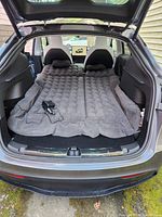 Black inflatable car air mattress set up flat in the trunk of an SUV with two pillows and inflator connected.
