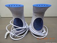 Photo showing a set of two identical portable garment steamers with blue and white color scheme, power cords coiled beside them.