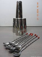Photo of four polished stainless steel Boston cocktail shakers stacked vertically showing their typical rounded shapes and smooth reflective surfaces, eight long stainless steel bar spoons with twisted handles and red tips laid out in front.