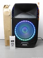 Front view of ION Total PA Extreme speaker powered on with multi-color LED woofer light display. Includes a tall cardboard box and an accessory container placed beside the speaker.
