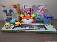 Front view of all items in the lot showing the Kiddo Lab Peekaboo Donkey plush toy in box, aquatic bath toys (yellow duck, red crab, blue hippo), electronic keyboard, water table pump in box, and 'Make It Real' spa kit.