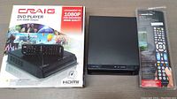 Craig DVD player with HDMI output next to its original box and Monster universal remote in packaging on a wooden surface.