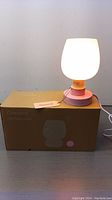 Photo of one lit LED table lamp atop its box, showing the product branding One Wish and LED Table Lamp text. Lamp has a pink base with a rounded white frosted shade, emitting warm light.
