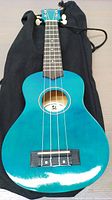 Top front view of blue ukulele showing body, soundhole, strings, and black carry bag beneath.