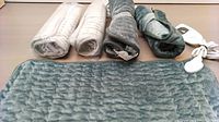 Five heating pads arranged side by side - three grey-blue quilted and two white rolled pads wrapped in plastic.