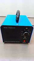 Blue rectangular ozone generator device with black front panel, timer dial labeled with minutes up to 180, and black carrying handle on top.