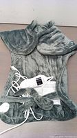 Gray plush heated back and shoulder wrap with attached cords and controller laying on flat surface.